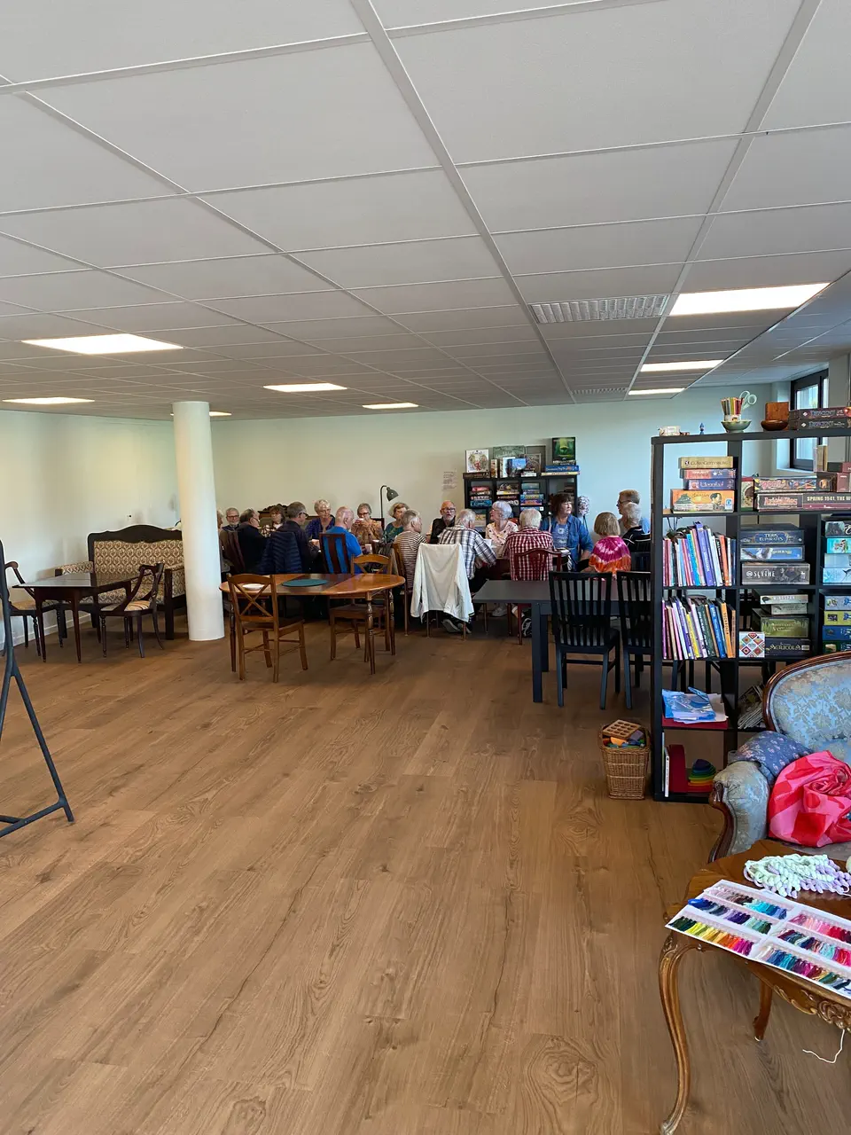 Caféets stora rum med långbord, gäster som fikar och spelhyllorna längs väggen